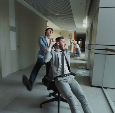 Two male colleagues having fun in an office corridor, with one seated on an office chair and the other playfully pushing him.
